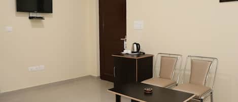 A room with a flat-screen TV mounted on the wall, a dark wooden table with a small tray, two metal-framed chairs with beige cushions, and a dark wooden door.