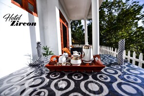 A tray with tea set and a teapot on a patterned table outside a building with a white fence and greenery in the background. - Zircon Ella (Ella)