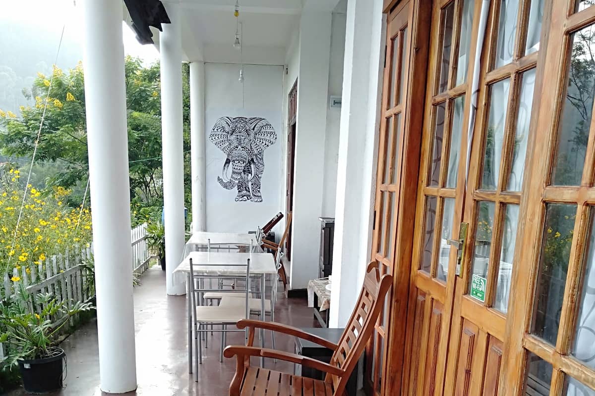 Un espacio al aire libre techado con sillas y mesa de madera, pilares blancos y un mural de un elefante.