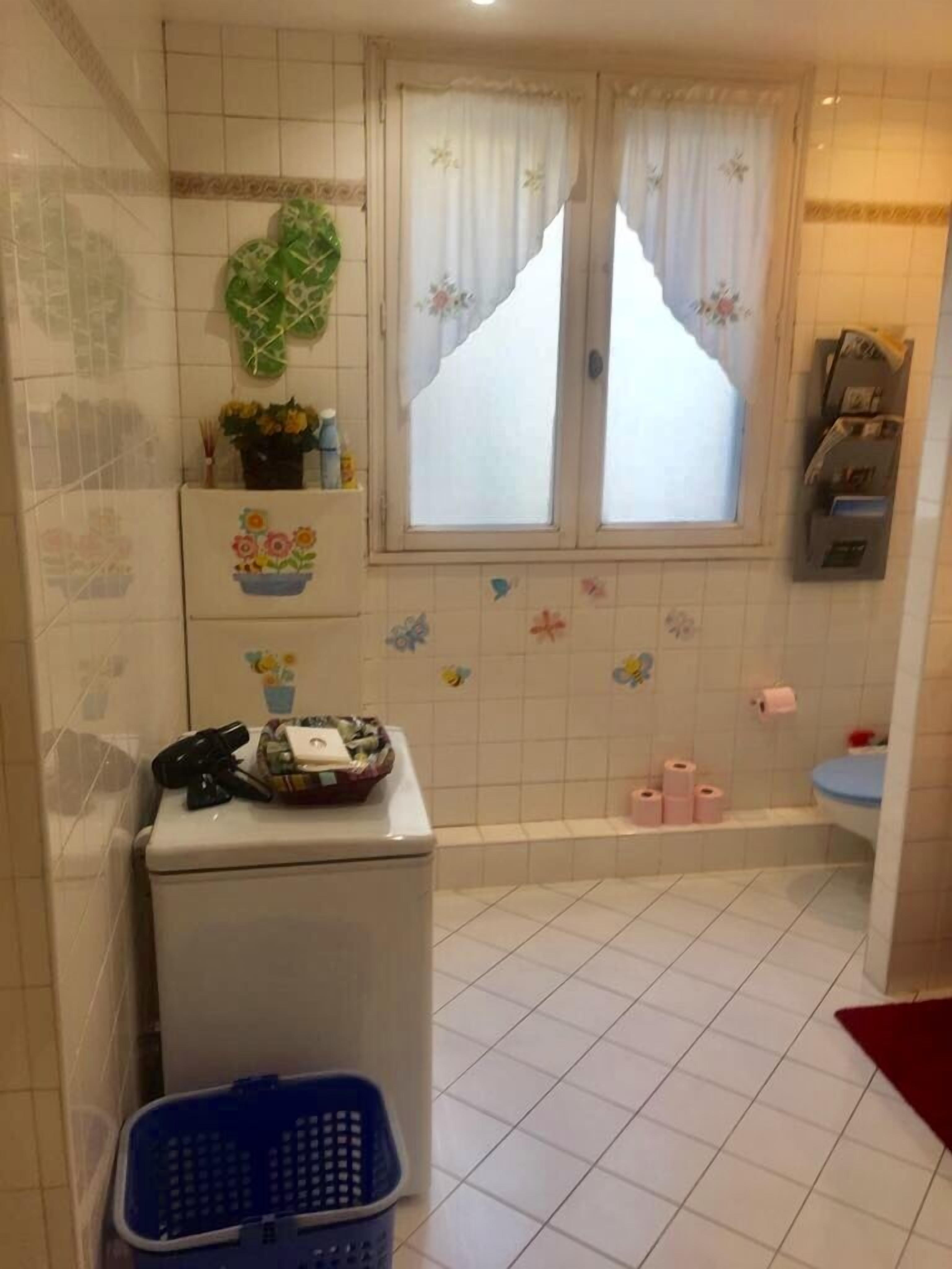 Un baño con inodoro, lavamanos, ventana con cortinas y un pequeño estante con artículos de aseo.