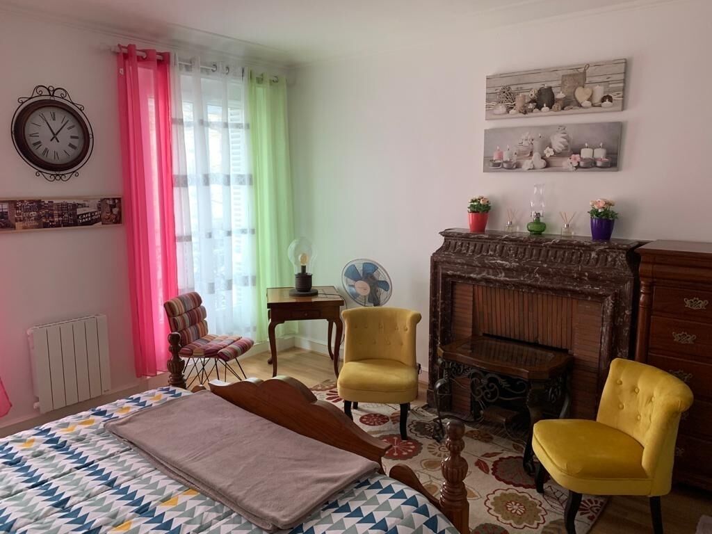 A bedroom with a bed, two yellow chairs, a wooden dresser, a fan, and a fireplace.
