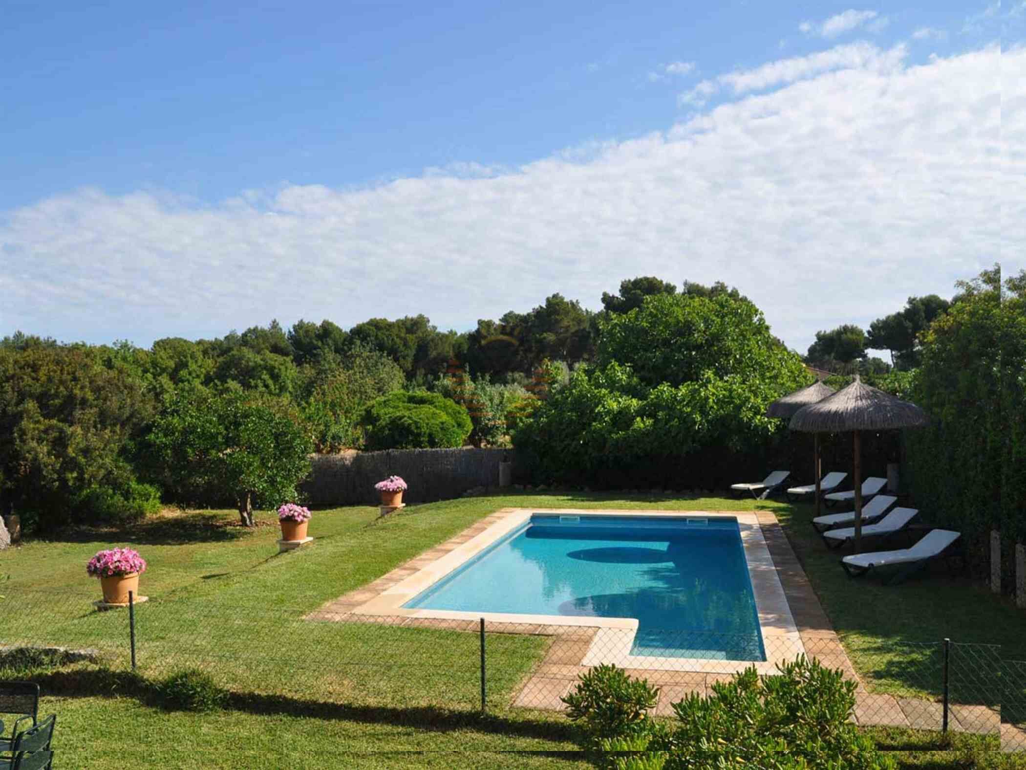 A rectangular swimming pool with a tiled border, surrounded by a well-maintained lawn and potted plants. There are lounge chairs and a small gazebo with a thatched roof in the background.