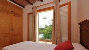 A bedroom with a bed, a red pillow, wooden wardrobe, and a view of a swimming pool and greenery. - Can Mostatxet, Country House in Can Picafort, Mallorca (Can Picafort)