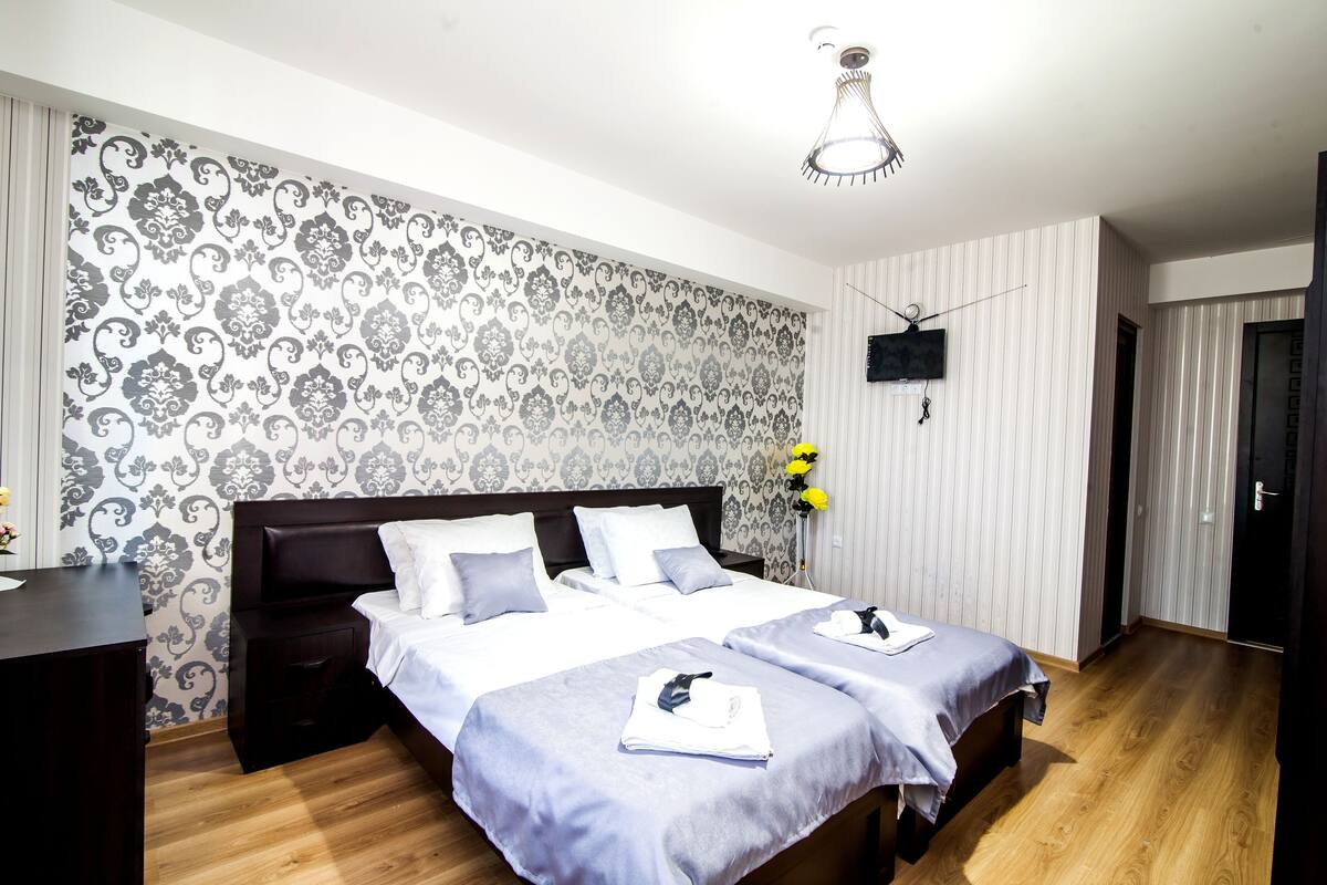 A hotel room with two beds, a wooden floor, a patterned wallpaper, a flat-screen TV, and a ceiling light fixture.