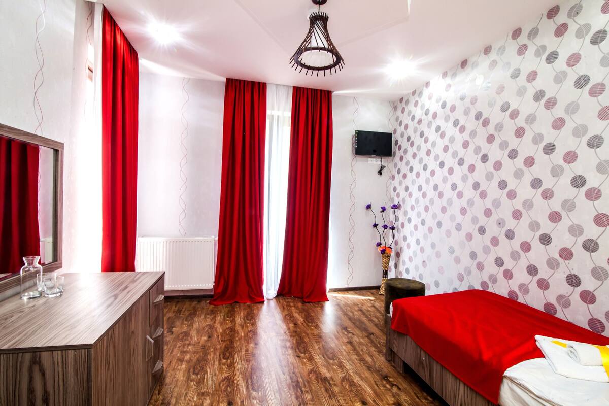 A hotel room with a wooden floor, a bed with a red blanket, a desk with a mirror, and a TV mounted on the wall.