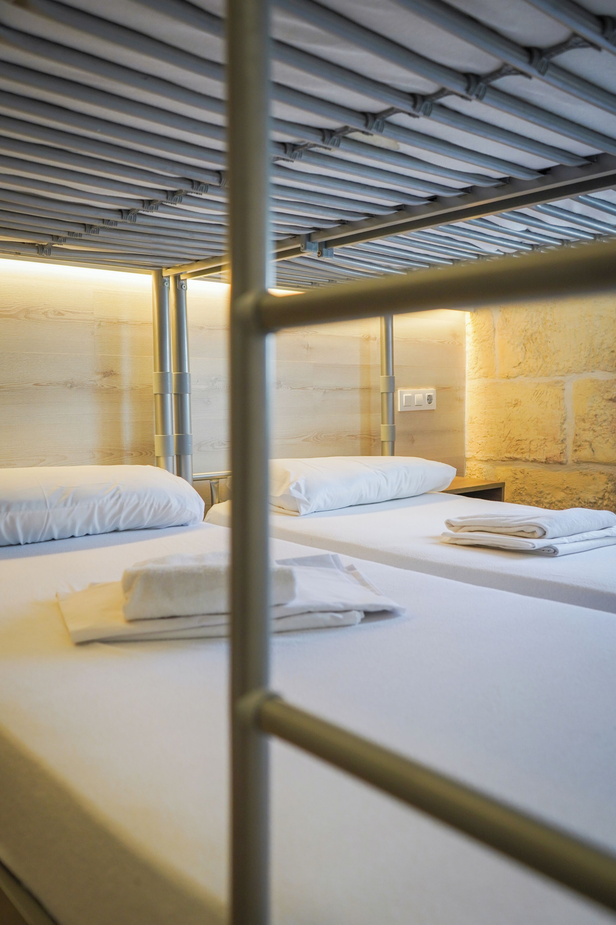 Habitación con literas de metal, ropa de cama blanca y un enchufe de pared.