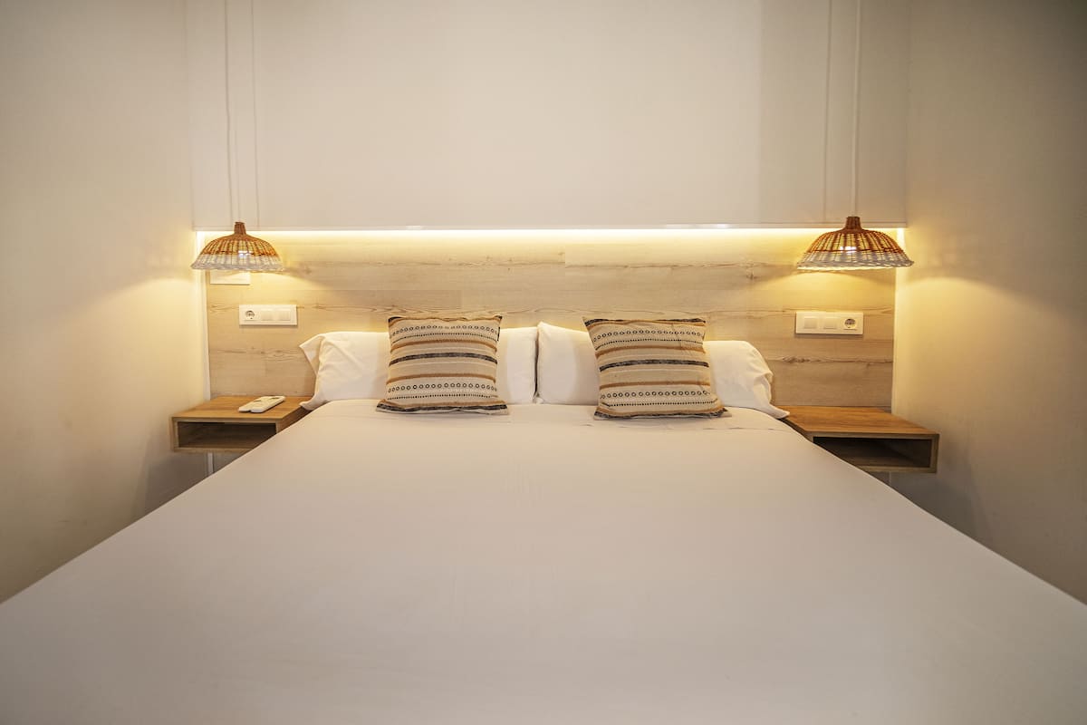 Una cama bien tendida con dos almohadas, dos mesitas de noche y dos lámparas fijadas a la pared.