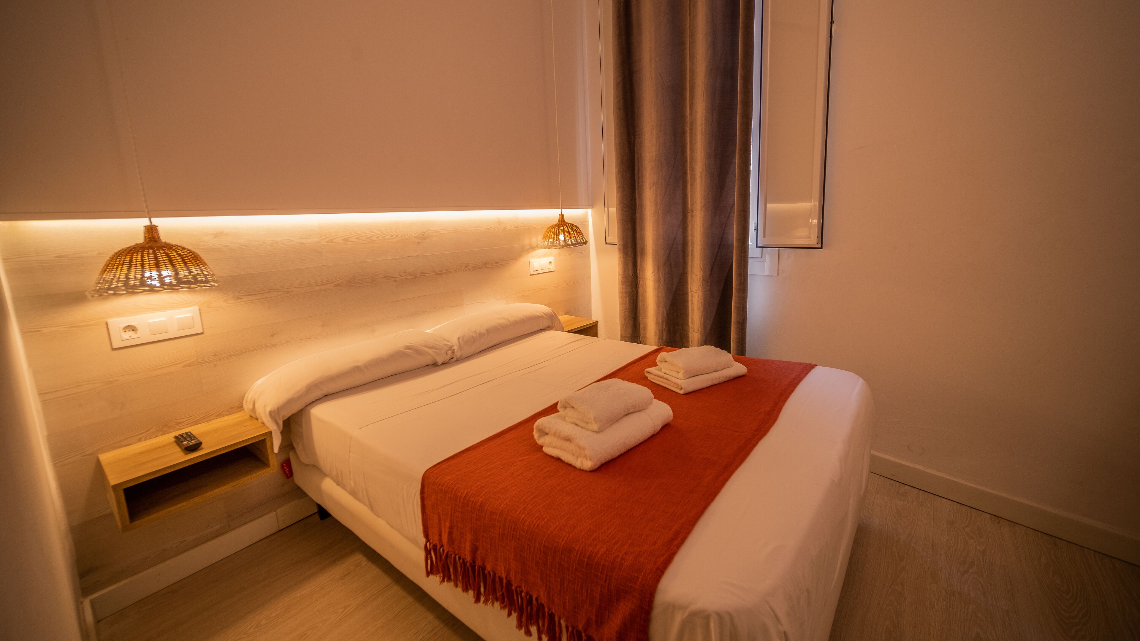 A neatly made bed with white linens, a red throw blanket, and two folded white towels. There is a bedside table with a lamp and a power outlet.