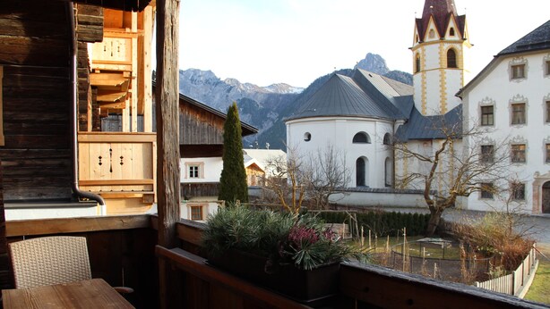 Un balcón de madera con una mesa y sillas, con vista a un patio que incluye una iglesia y montañas al fondo.