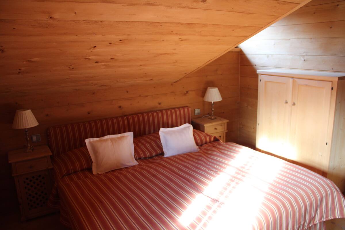 Un dormitorio con techo inclinado, una cama con ropa de cama a rayas, una mesita de noche con lámpara y un armario.