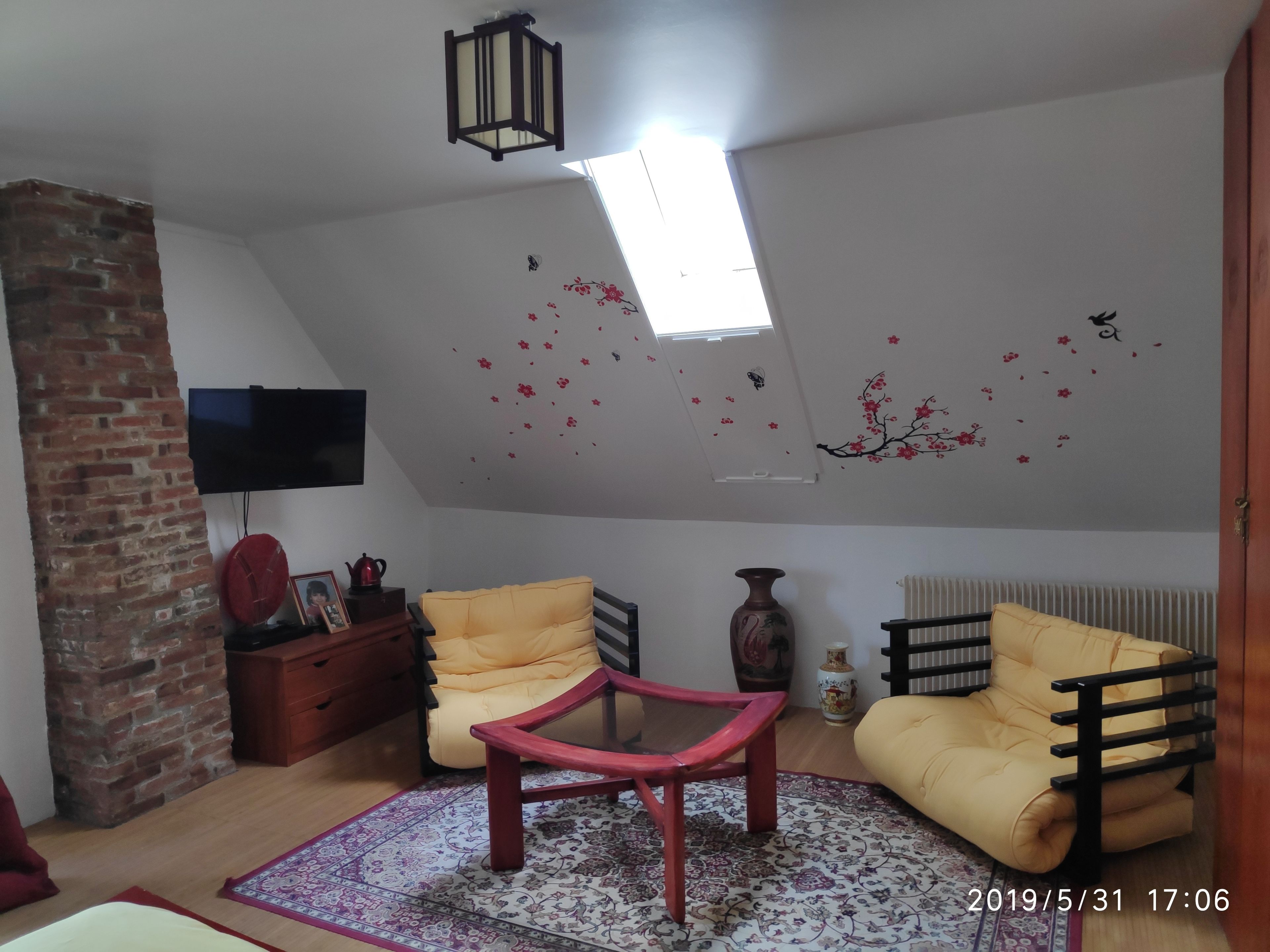 Una habitación estilo loft con techo inclinado, pared de ladrillo, televisor montado en la pared, dos sillas amarillas, una mesa roja y una alfombra estampada.