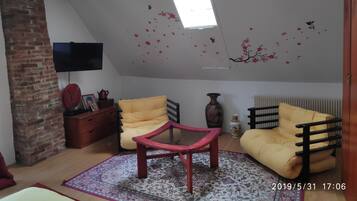 Una habitación estilo loft con techo inclinado, pared de ladrillo, televisor montado en la pared, dos sillas amarillas, una mesa roja y una alfombra estampada.