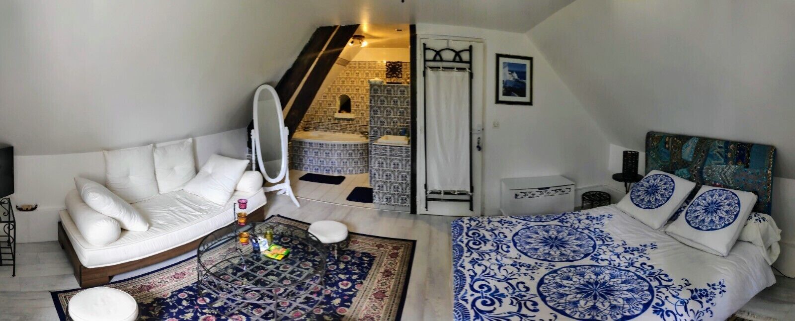 Une chambre avec un canapé blanc, un lit avec une literie à motifs bleus et blancs, une petite table ronde avec un miroir et un élément décoratif mural.