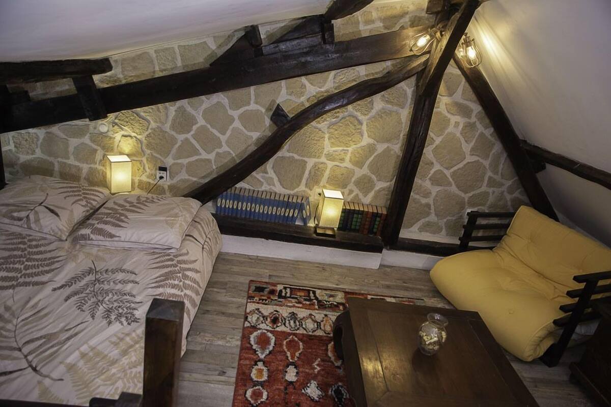 Een gezellige kamer met een bed, een gele fauteuil, een houten tafel en een kamerbreed tapijt. Er zijn twee wandlampen en een boekenplank met boeken.