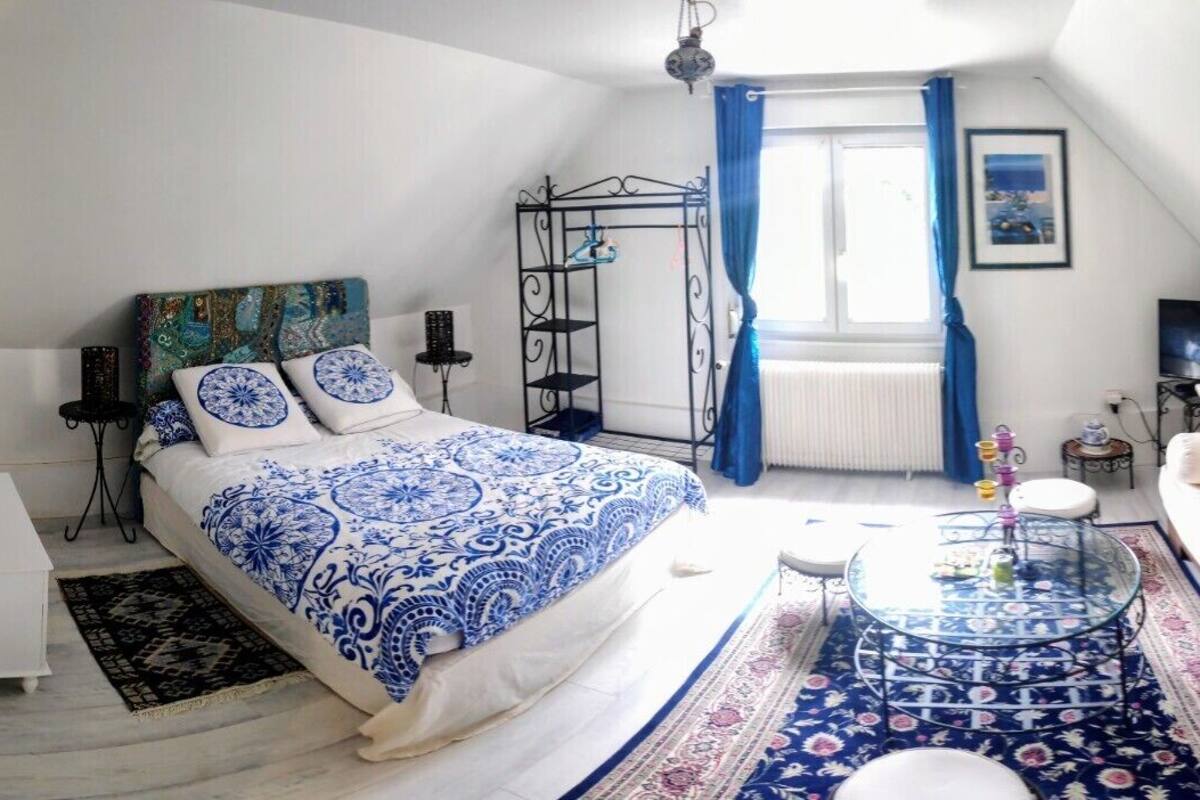 Een slaapkamer met een bed, een sofa, een glazen bijzettafeltje, een radiator, een raam met gordijnen en een schilderij aan de muur.