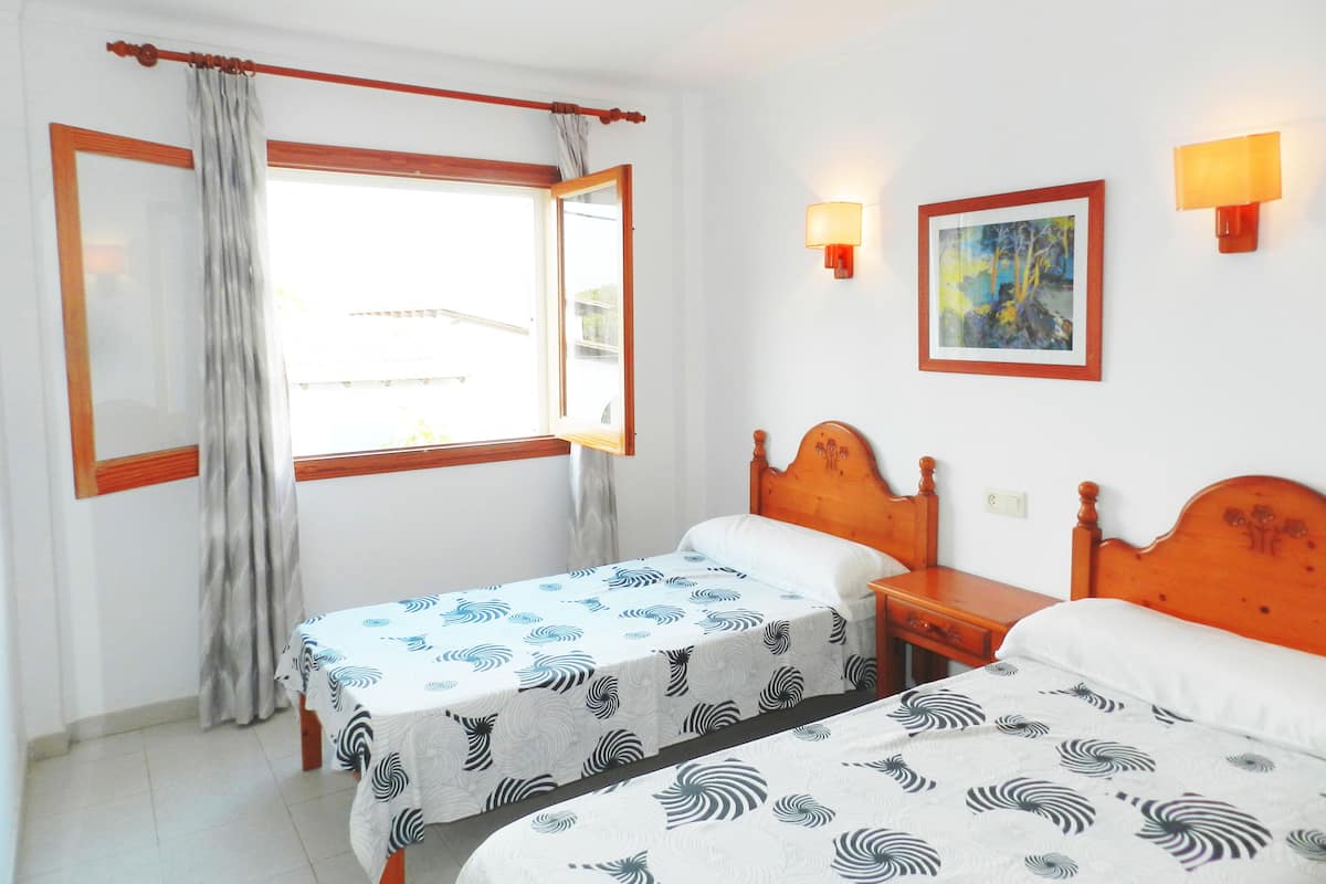 Une chambre avec deux lits, une fenêtre avec des rideaux, une lampe fixée au mur et un tableau encadré.