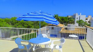 Een terras met een blauw-wit gestreepte parasol, witte plastic stoelen en een kleine ronde tafel.
