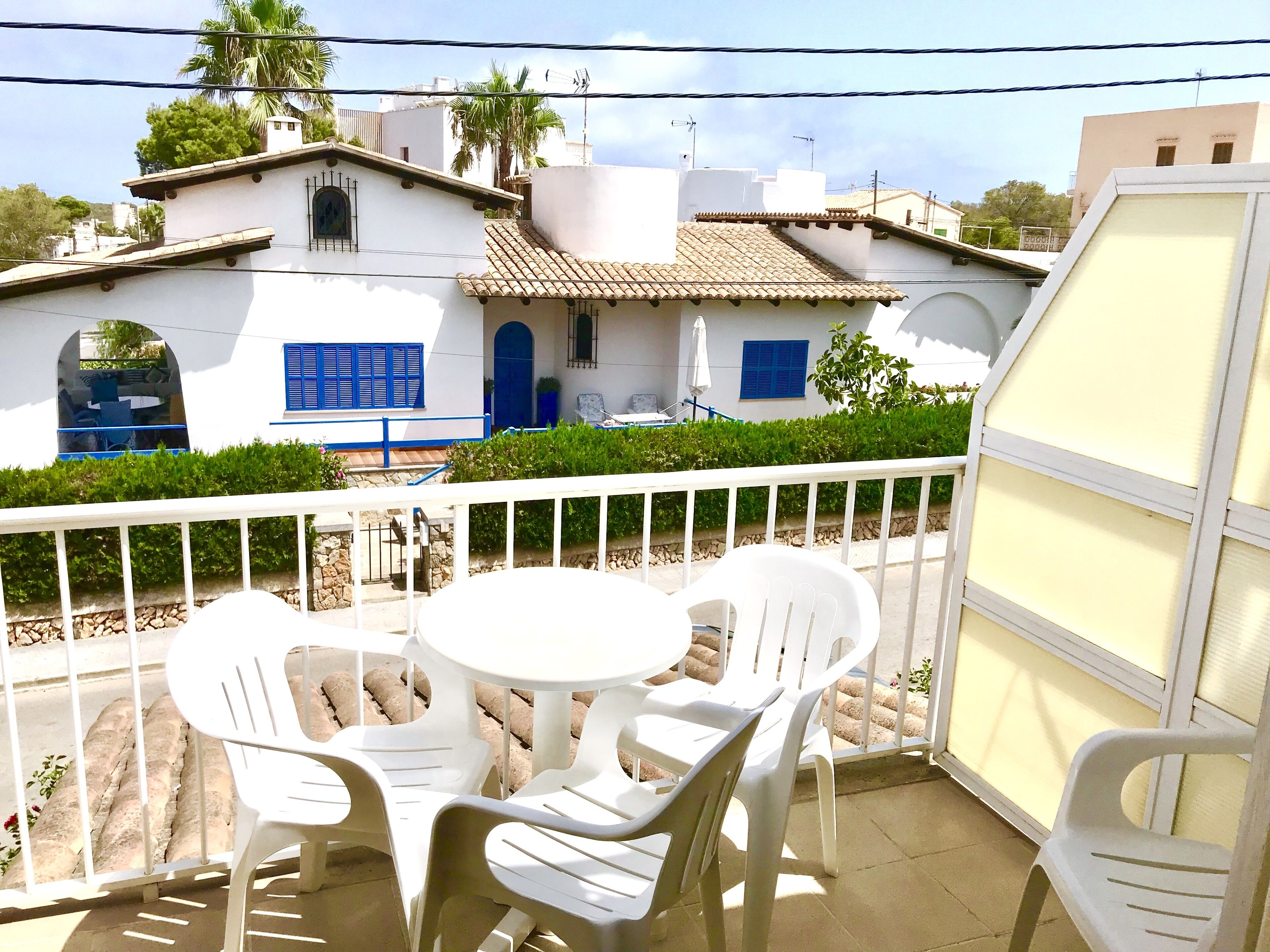 A balcony with white plastic chairs and a table, overlooking a white building with blue shutters and a tiled roof.