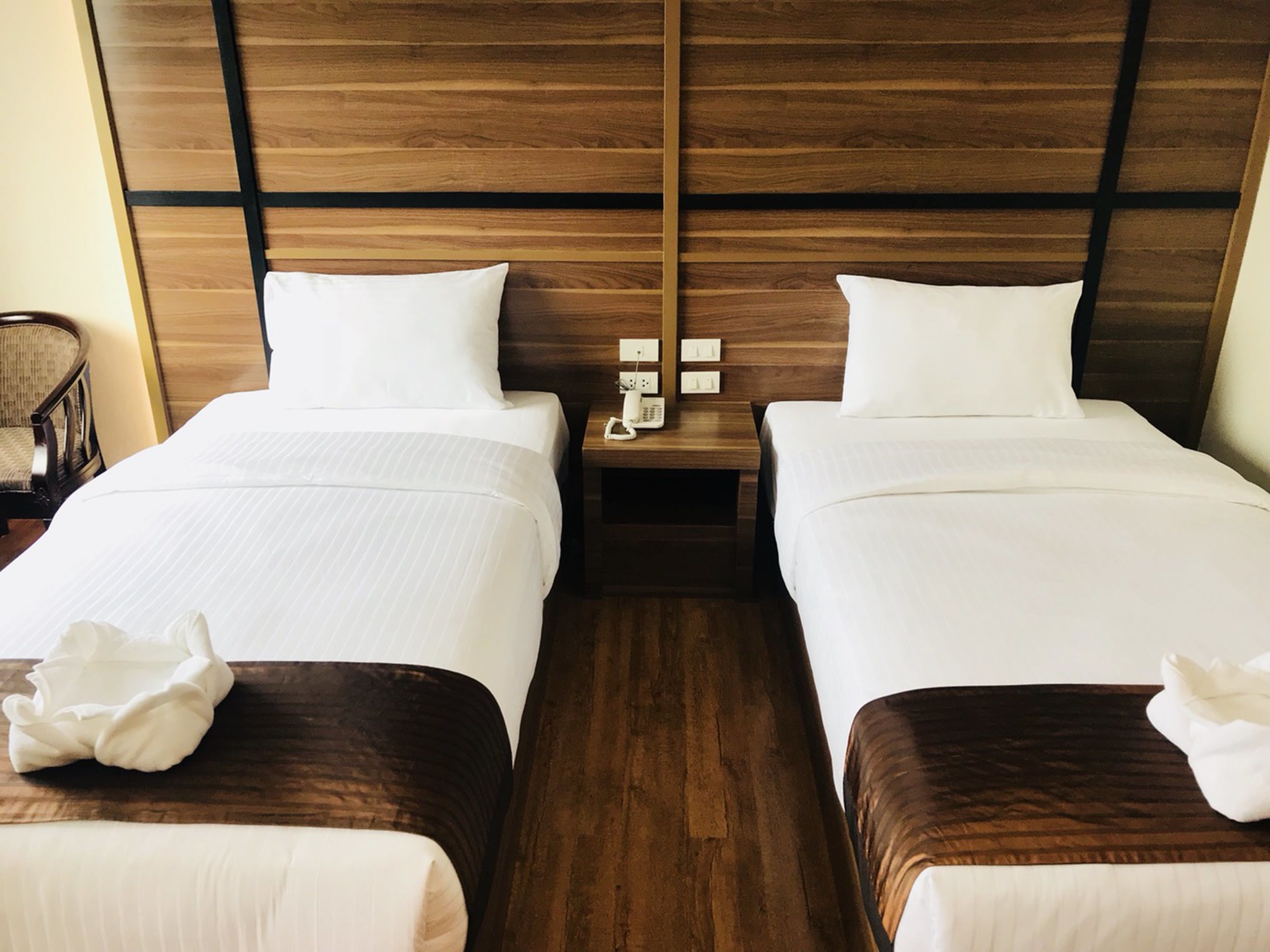 A hotel room with two beds, wooden headboards, a nightstand with a phone, and a chair.