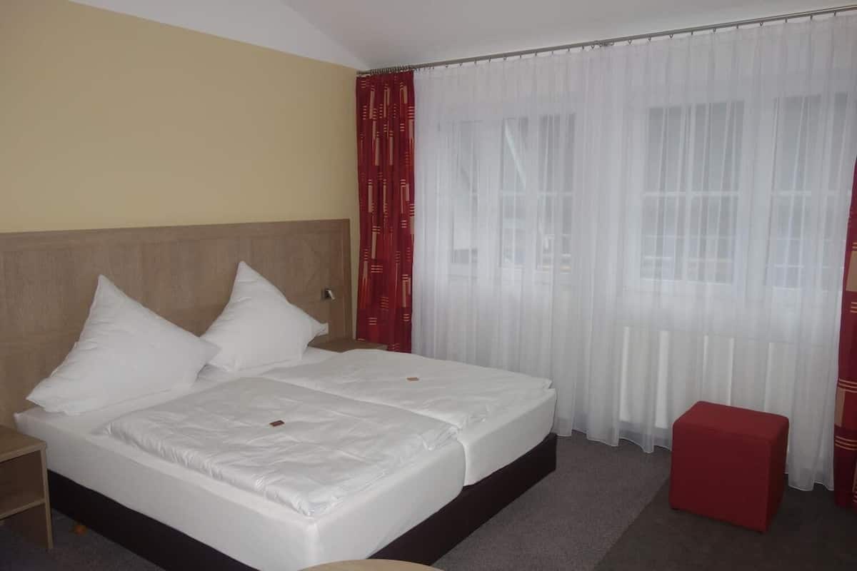 A hotel room with a double bed, a wooden headboard, a red ottoman, and sheer curtains.