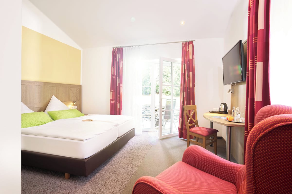 A hotel room with a bed, a TV, a chair, and a table with a kettle and glasses.