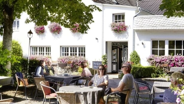 An outdoor seating area with tables and chairs, surrounded by lush greenery and a white building with hanging flower baskets.