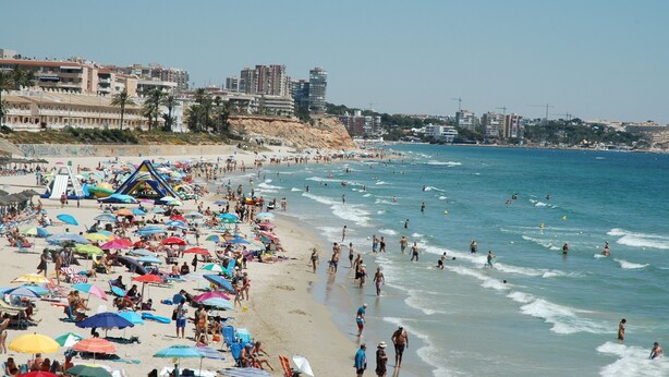 Ein überfüllter Strand mit Sonnenschirmen, Menschen und Gebäuden im Hintergrund.