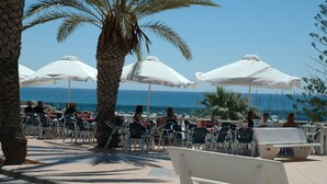 Sitzgelegenheiten im Freien mit Sonnenschirmen und Tischen, Palmen und Blick auf das Meer.
