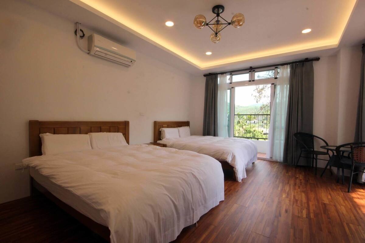 Ein Hotelzimmer mit zwei Betten, einem Deckenventilator, einer Klimaanlage, einem Essbereich mit Tisch und Stühlen sowie einem großen Fenster mit Vorhängen.