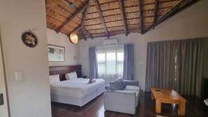 A hotel room with a bed, a sofa, a wooden table, and a large window with curtains. - Qabuleka bnb (Nongoma)