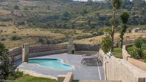 A swimming pool with a stone wall and lounge chairs, surrounded by a landscaped garden and a hilly backdrop. - Qabuleka bnb (Nongoma)