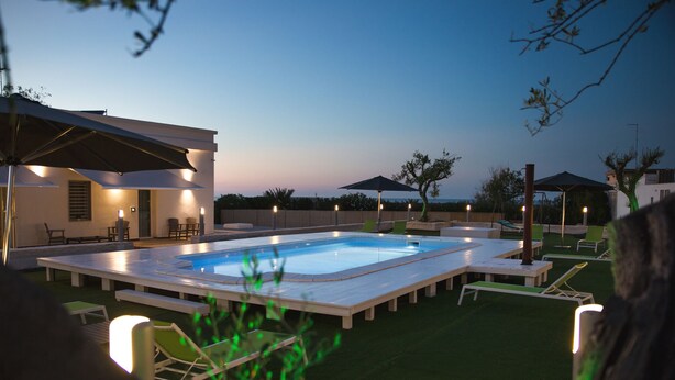 Piscina retangular com um deck ao redor, mobiliário de exterior e iluminação ambiente.