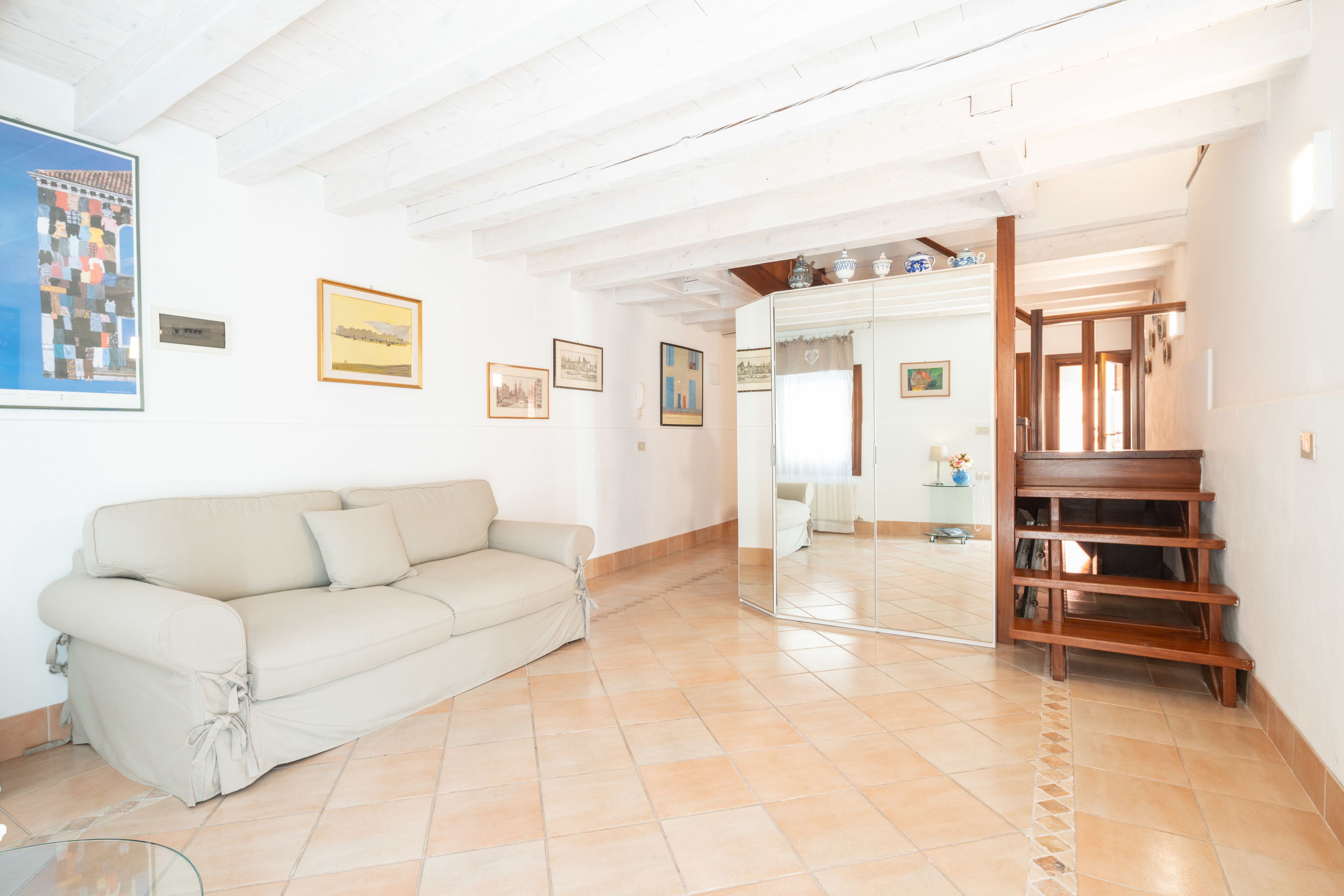 A spacious living area with a beige sofa, tiled flooring, and a wooden bookshelf.