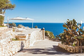 Un chemin de pierre mène à une terrasse agrémentée de chaises longues blanches et d’un grand parasol, d’où l’on peut admirer une vue sur la côte.