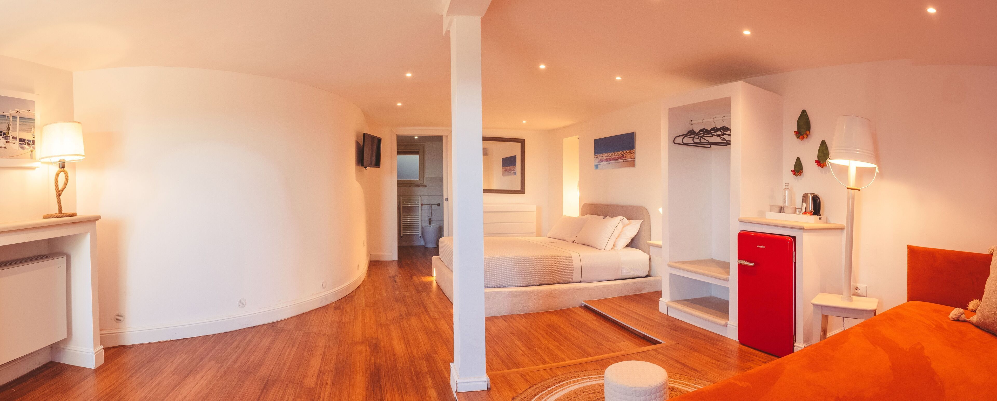 Une chambre d’hôtel moderne avec un mur blanc incurvé, un plancher en bois, un lit avec une literie blanche, un mini-bar rouge, une petite table et un téléviseur fixé au mur.