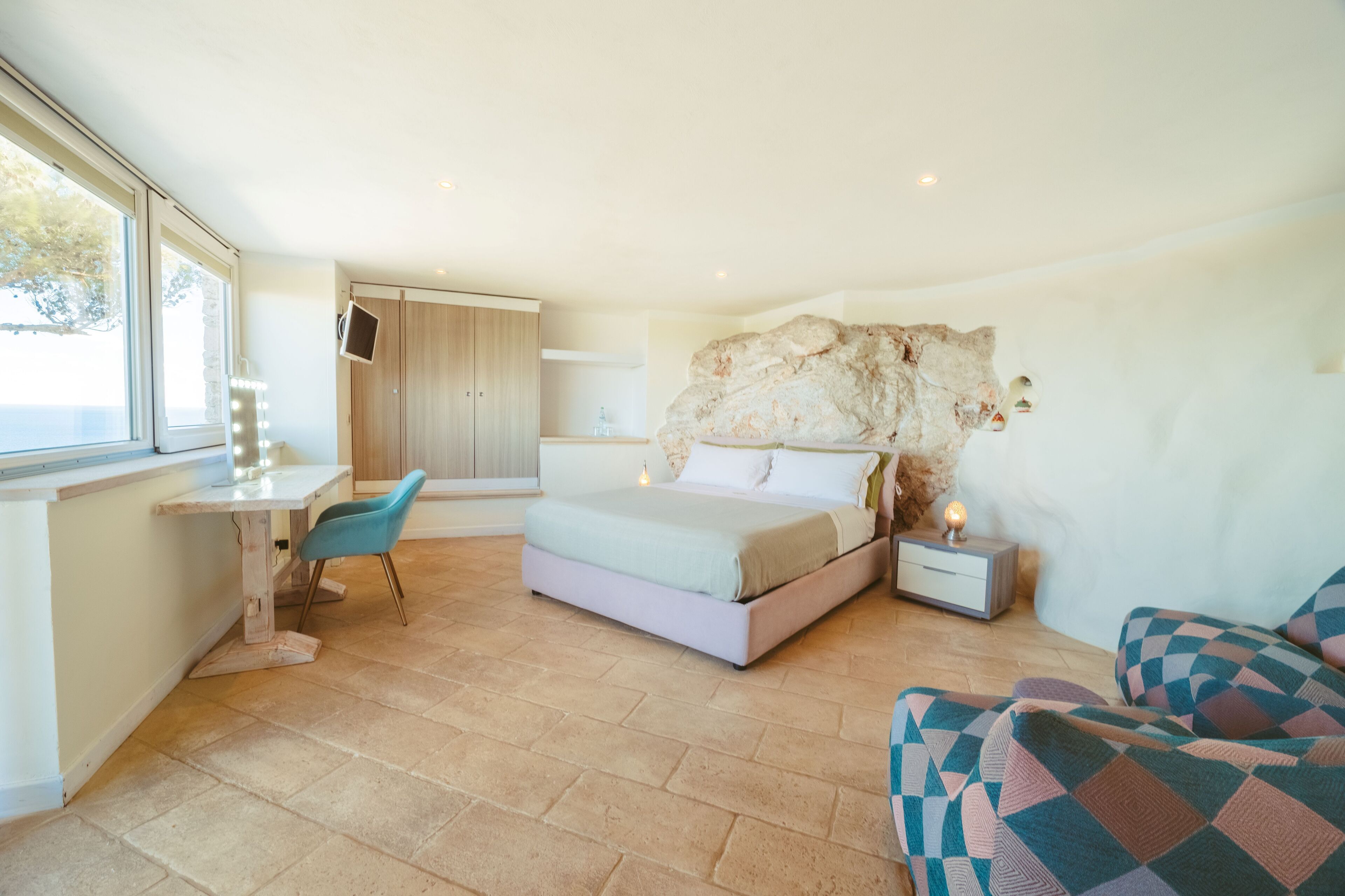 A bedroom with a bed, a desk, a chair, a nightstand, and a stone wall.