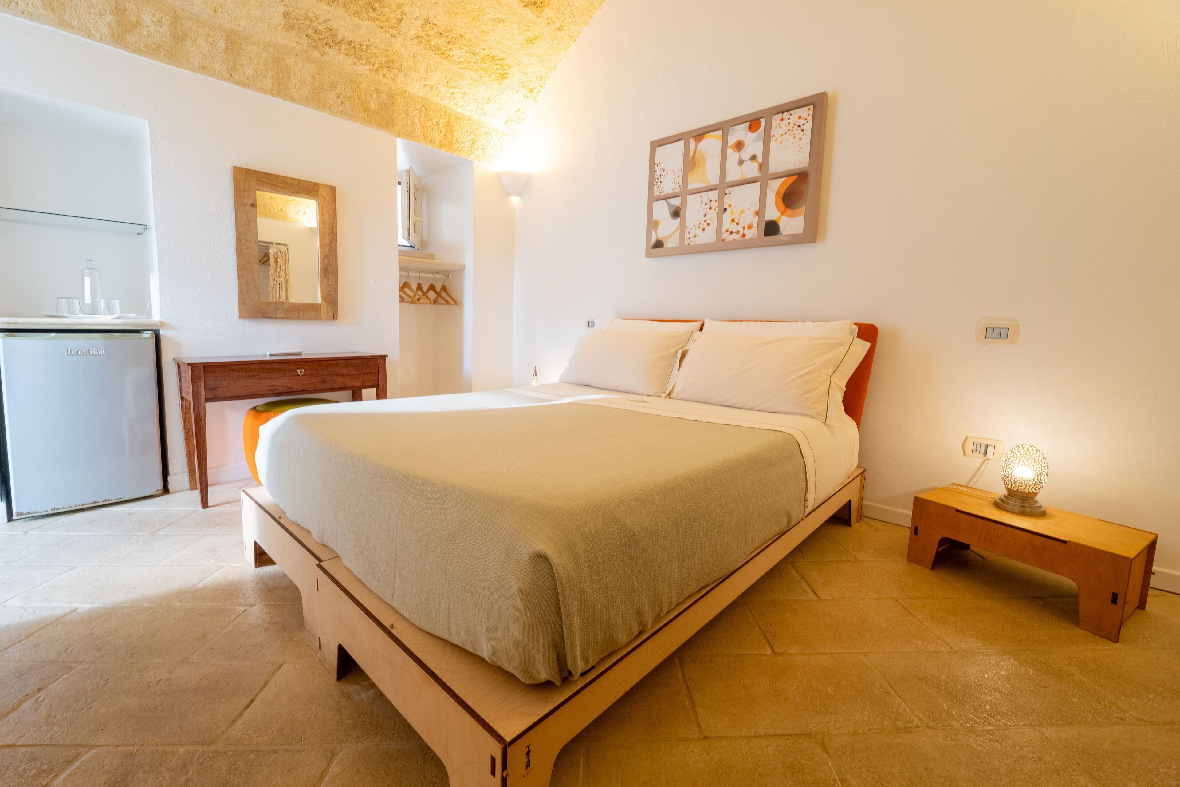 A bedroom with a bed, a wooden nightstand, a desk, and a mirror.