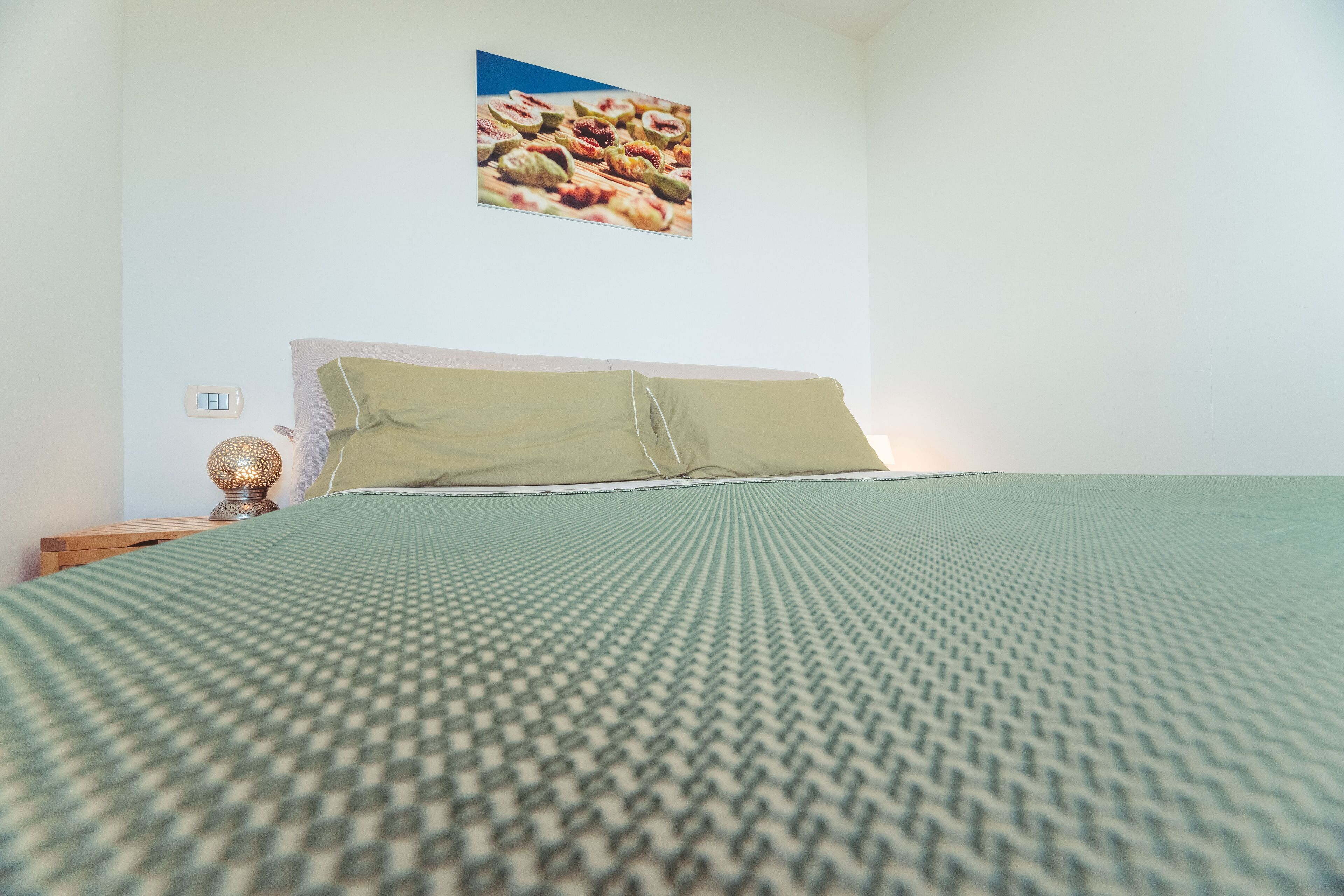 A neatly made bed with a patterned bedspread, a pillow, and a wall-mounted picture of fruits.