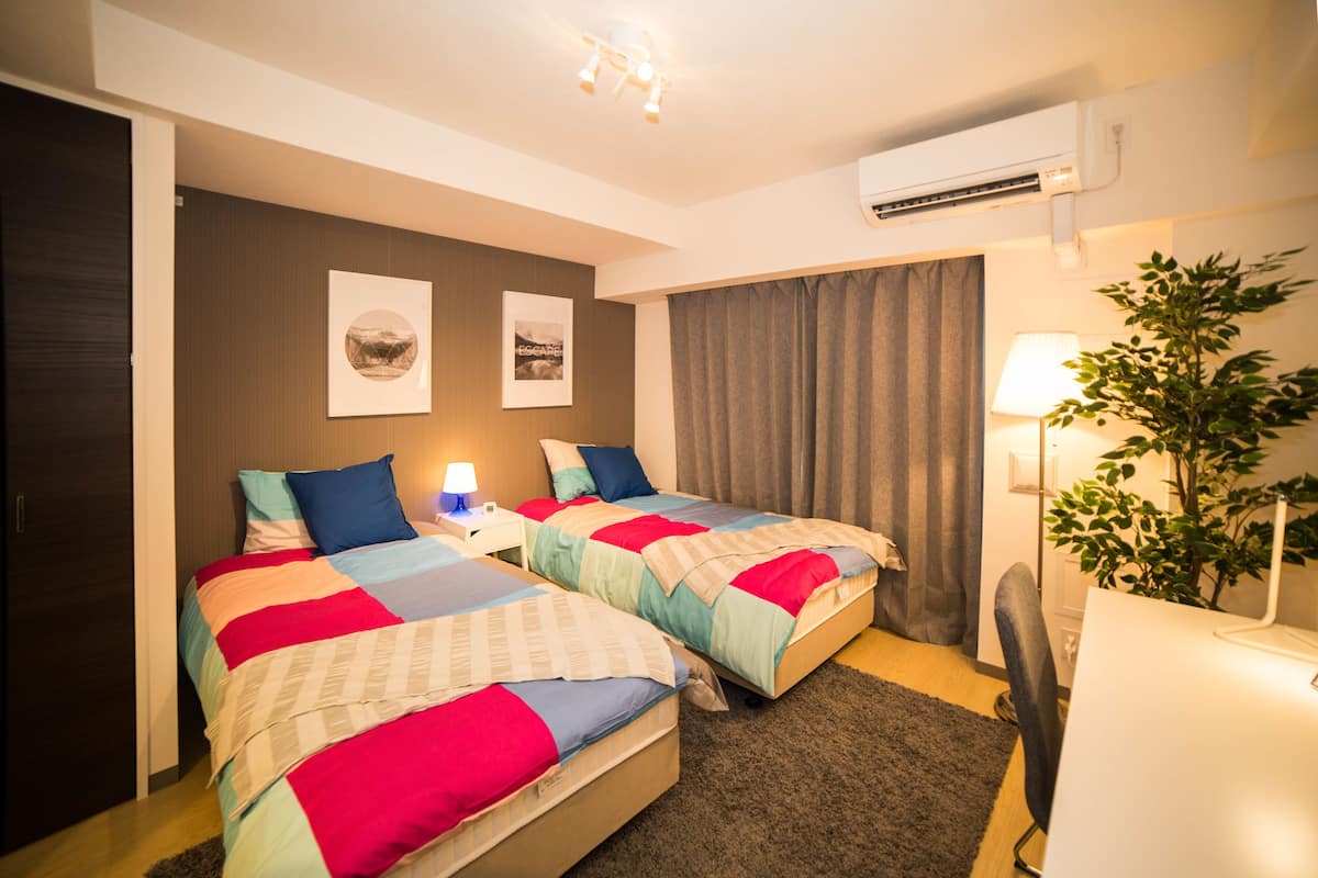 Habitación de hotel con dos camas, un escritorio, una silla, una planta y un aire acondicionado.