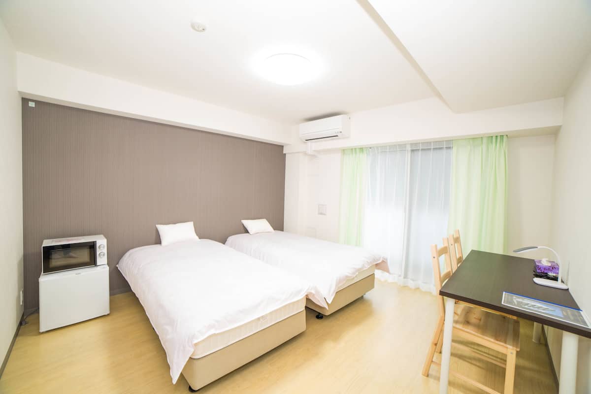 Habitación de hotel con dos camas, una kitchenette pequeña, una mesa de comedor y un ventanal con cortinas.