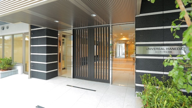 Entrada de um edifício moderno com fachada listrada em preto e branco, porta de vidro e uma placa com a inscrição 'UNIVERSAL HANEDA'.