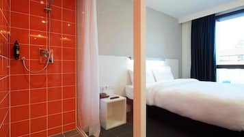 Hotelkamer met een douchegedeelte met rode tegels, een witte nachtkastje met lamp en een groot bed met witte lakens.