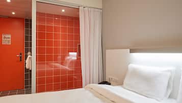 Une chambre d’hôtel avec un lit, une salle de bain aux carreaux rouges, un rideau blanc et une tête de lit blanche.