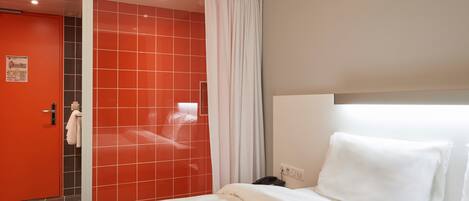 A hotel room with a bed, a bathroom with red tiles, a white curtain, and a white headboard.