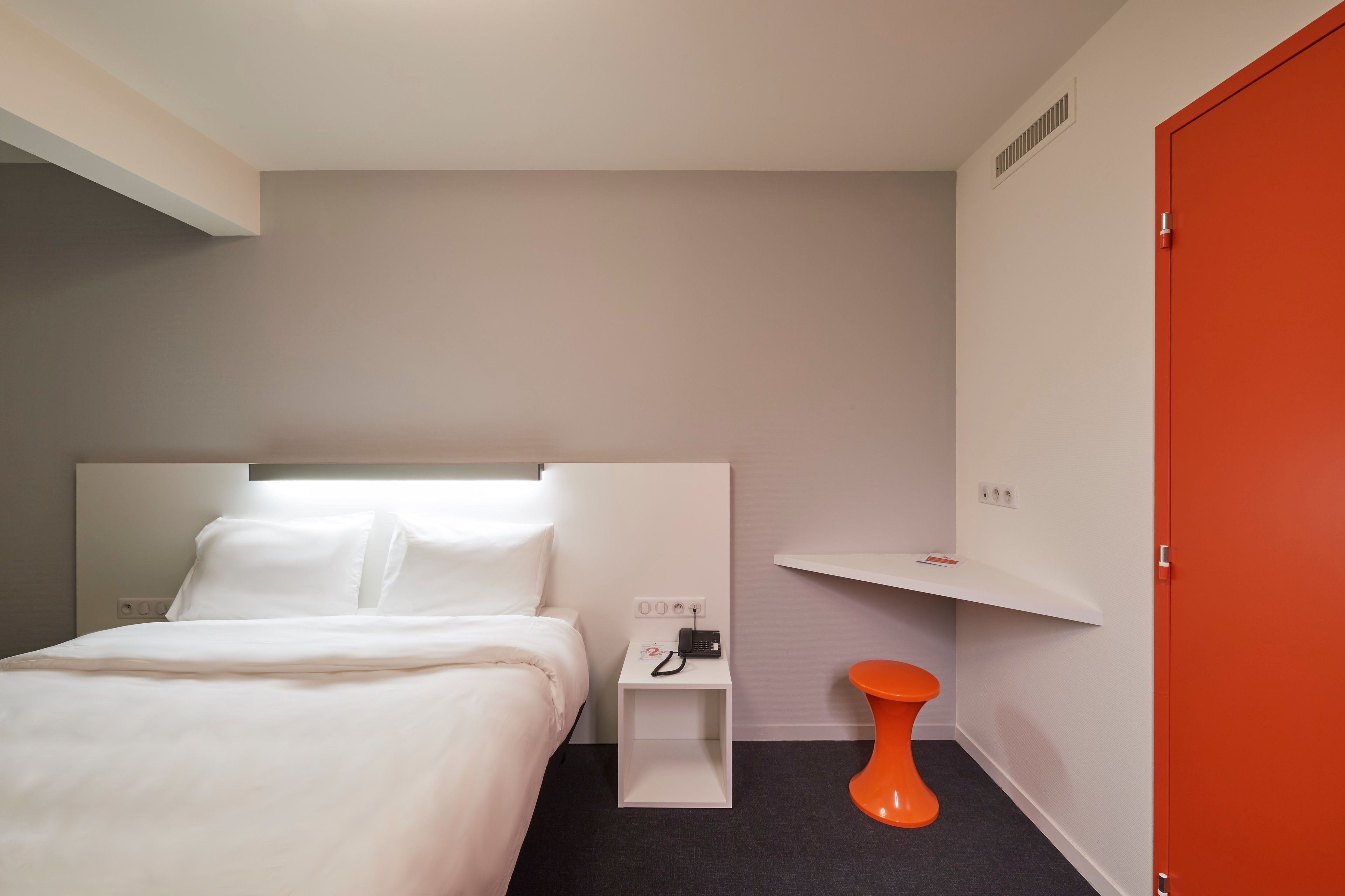 Een hotelkamer met een bed, een nachtkastje met een telefoon, een oranje kruk en een rode deur.