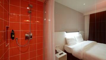 A hotel room with a red tiled wall, a shower, a bed with white bedding, a nightstand, and a small table with a remote control.