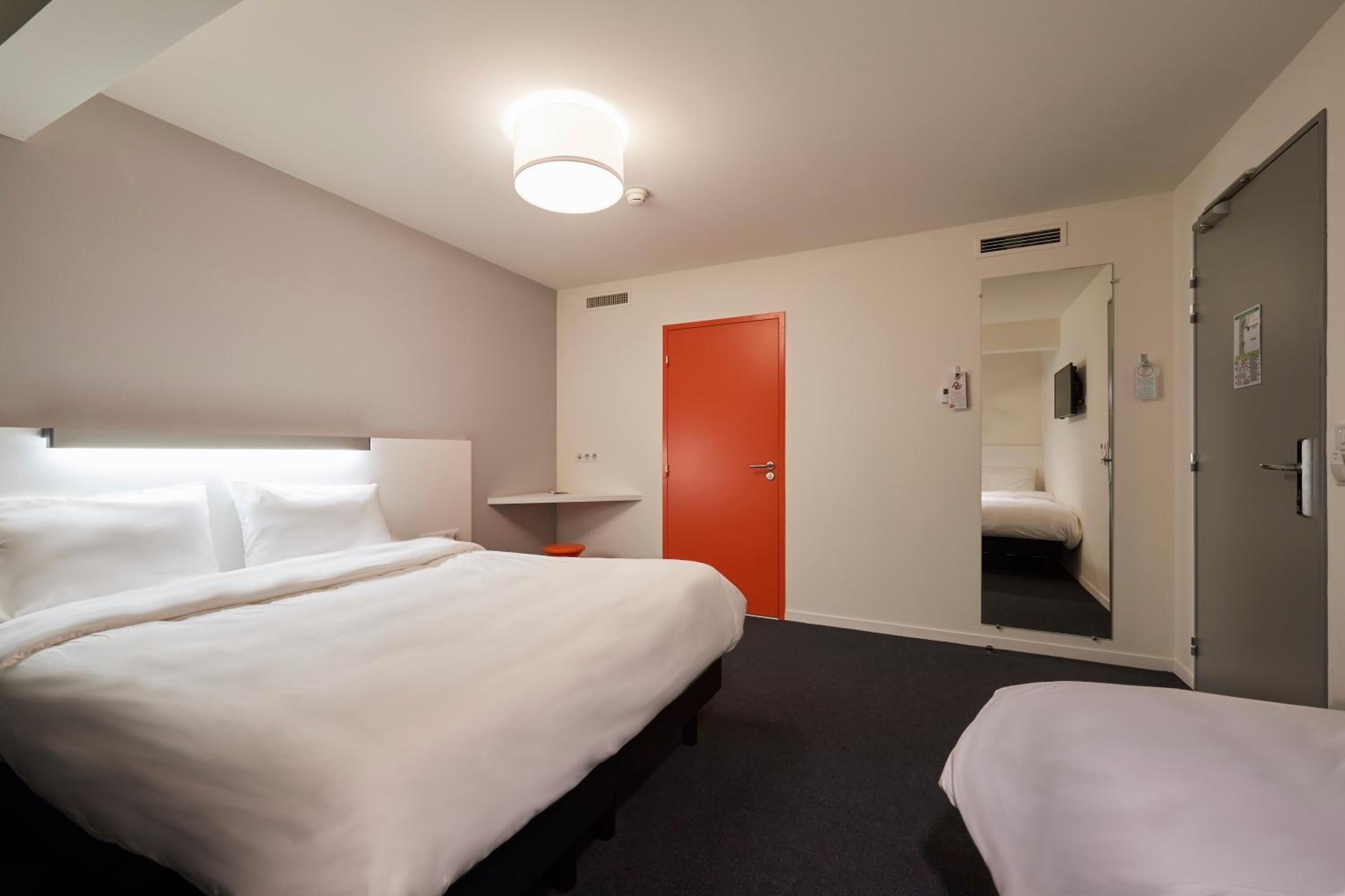 Een hotelkamer met een groot bed, een nachtkastje, een rode deur en een spiegel die de kamer weerspiegelt.