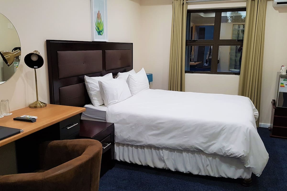 Ein Hotelzimmer mit Bett, Schreibtisch mit Stuhl, Spiegel, Nachttisch und einem Fenster mit Vorhängen.