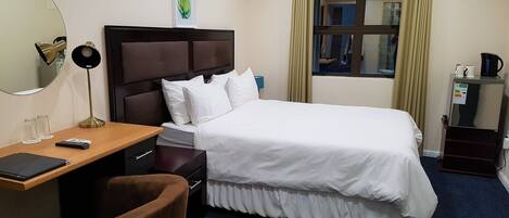 A hotel room with a bed, a desk with a chair, a mirror, a nightstand, and a window with curtains.