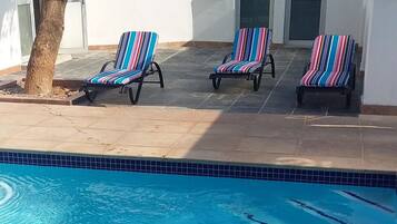 A swimming pool with three striped lounge chairs and a tree.